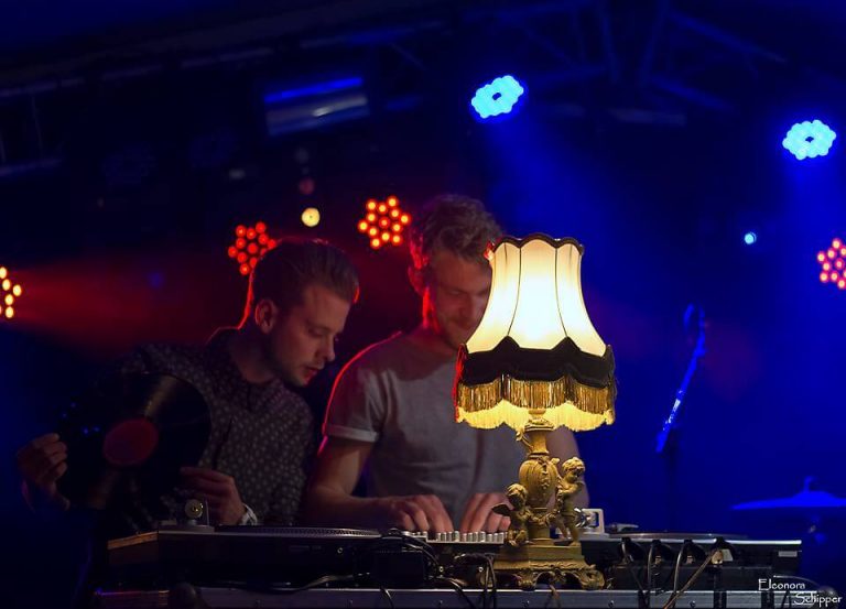 Twee DJ’s achter vinyl draaitafels met een schemerlamp en gekleurde verlichting