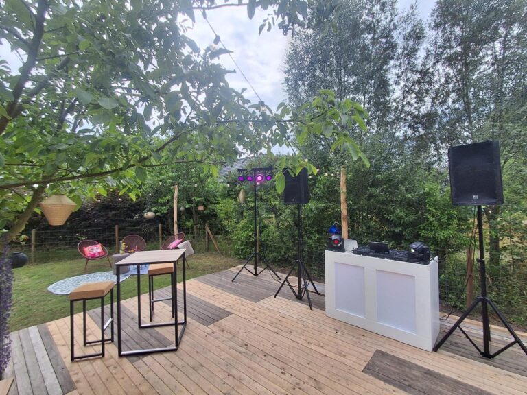 Witte DJ-booth met speakers en feestverlichting op een houten terras tussen het groen