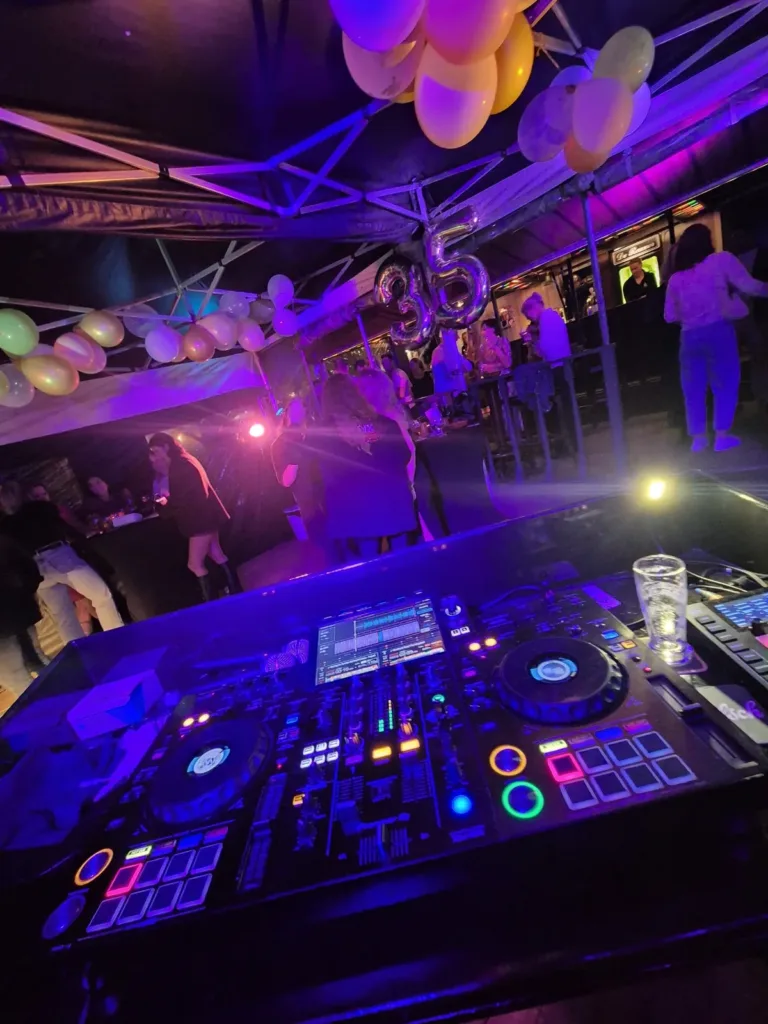 Uitzicht over een verlichte DJ controller naar de dansvloer met paarse feestverlichting