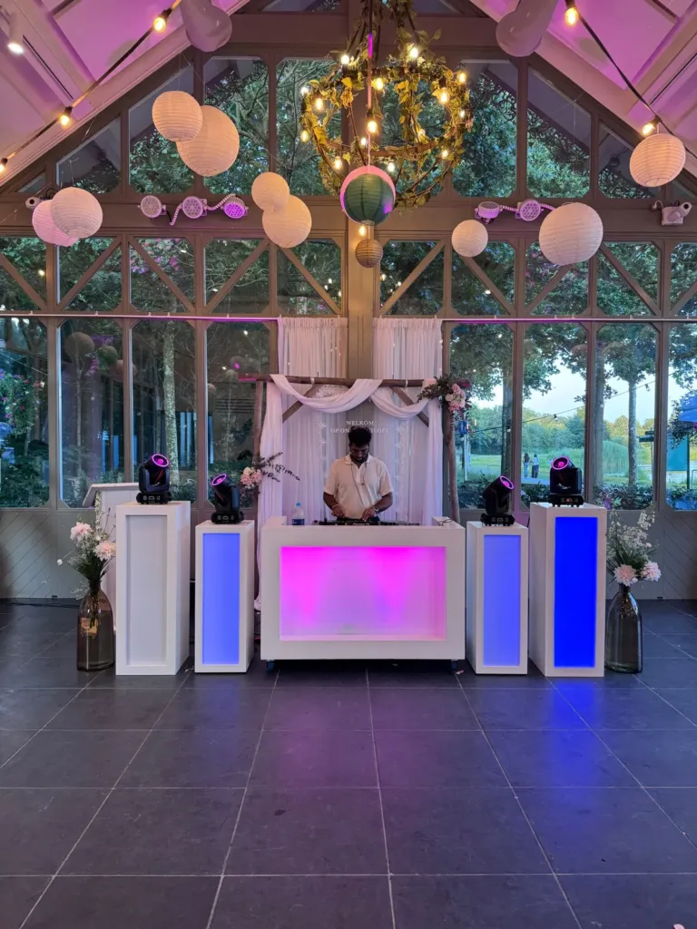 Witte DJ booth met kleurrijke lichtzuilen in een glazen kas met lampionnen en kroonluchter