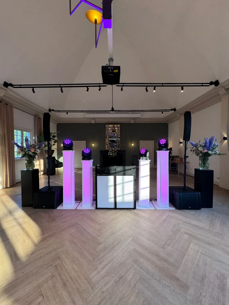 Witte DJ booth met paarse lichtzuilen en speakers in een lichte zaal met houten vloer