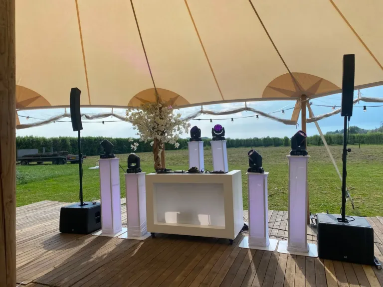 Brede witte DJ booth met vier lichtzuilen en speakers onder een stretchtent in het groen